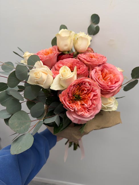A bouquet of fresh flowers featuring large, ruffled pink ranunculus and delicate white roses, accented with green eucalyptus leaves. The arrangement is wrapped in light brown paper with a subtle pink 