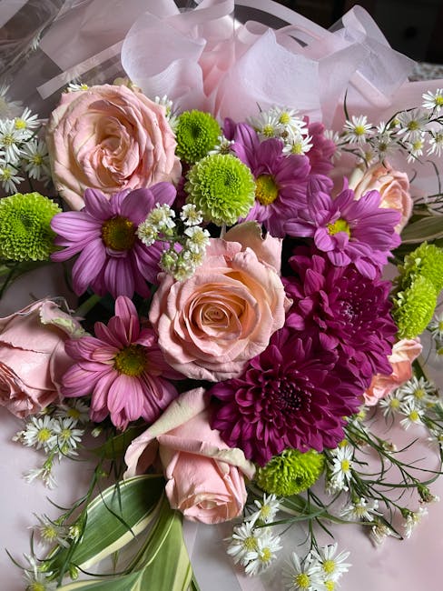 A vibrant bouquet featuring large blush pink roses with tightly packed petals, surrounded by bright purple daisies with yellow centers, and small white daisies with yellow centers. Interspersed among 