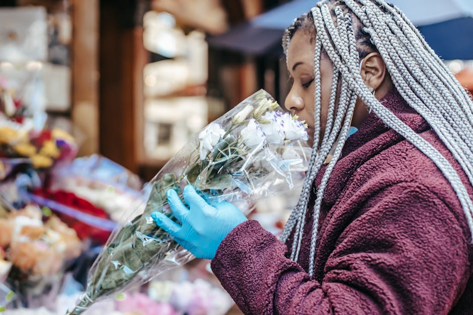 A woman at an outdoor flower market holding a bouquet of white roses wrapped in clear plastic, wearing a maroon fleece jacket and blue disposable gloves, with her face close to the flowers, examining 