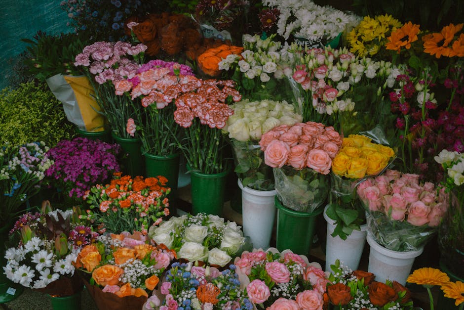 A vibrant display of fresh flowers in various colors and arrangements, including pink and peach roses, white and purple daisies, orange carnations, and pink alstroemerias, all neatly organized in gree