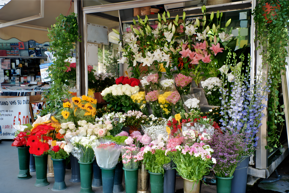 A vibrant flower display outside a floristry shop, featuring a wide range of fresh flowers arranged in various vases and buckets. Prominent blooms include white lilies with large, trumpet-shaped petal